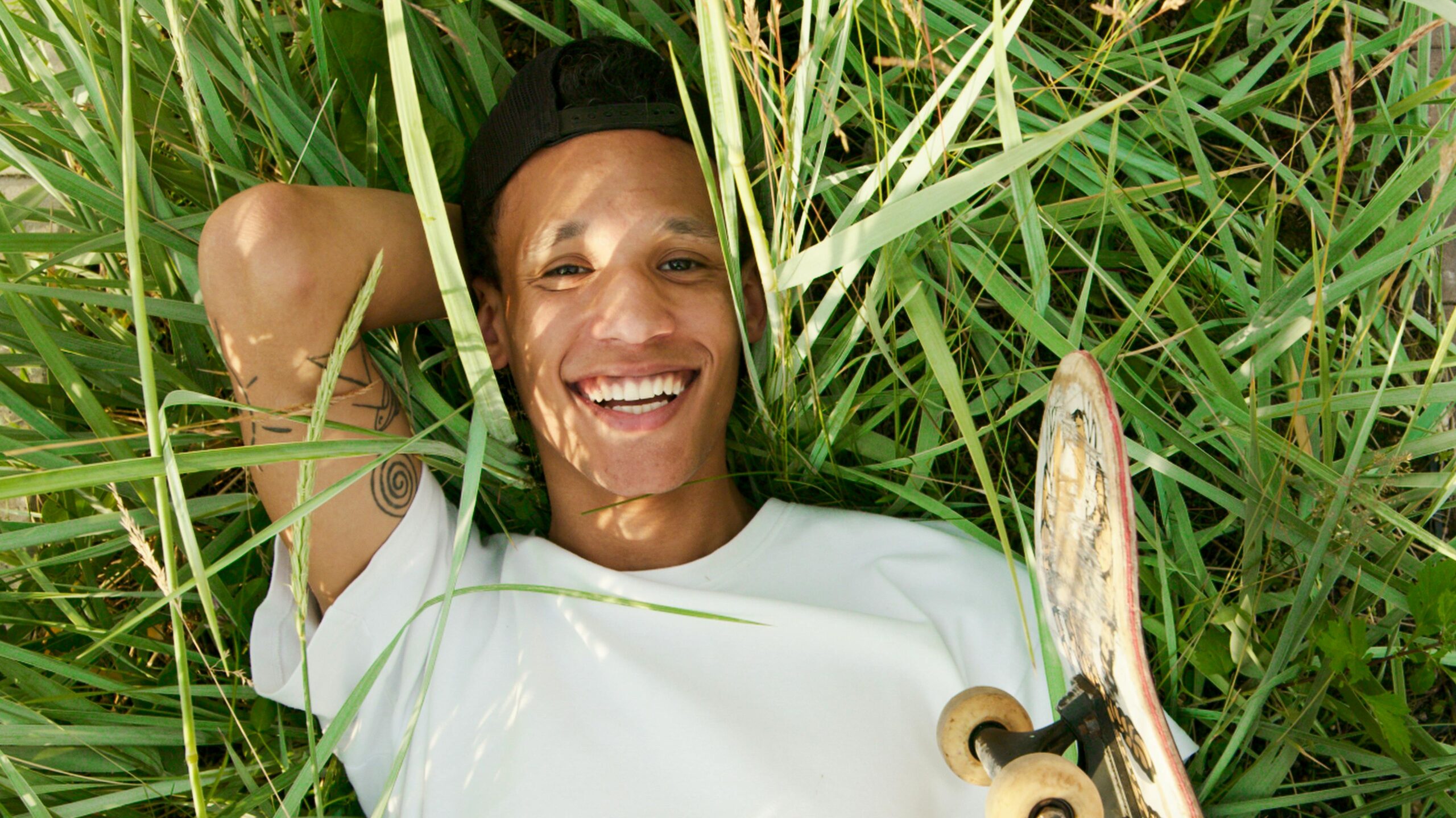 A cheerful young skateboarder lying in the grass on a sunny day, smiling with a skateboard beside him.
